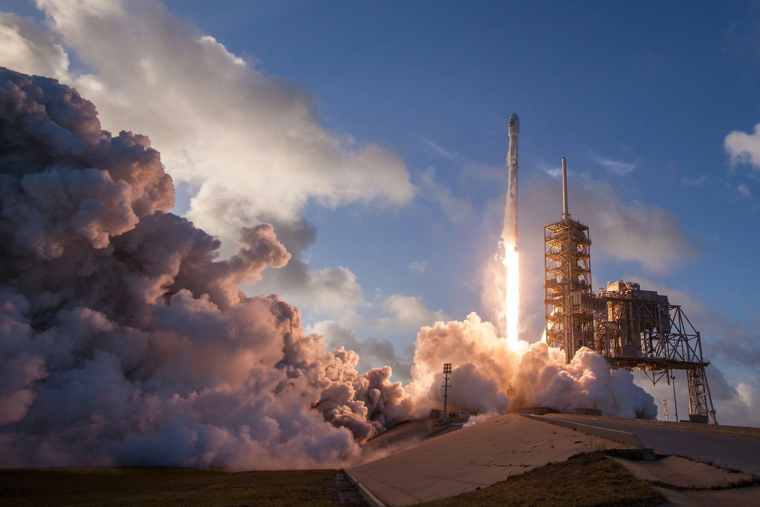 Rocket ship that is taking off surrounded by smoke from the blast. The ship is just off the ground and has a blue sky in the background.