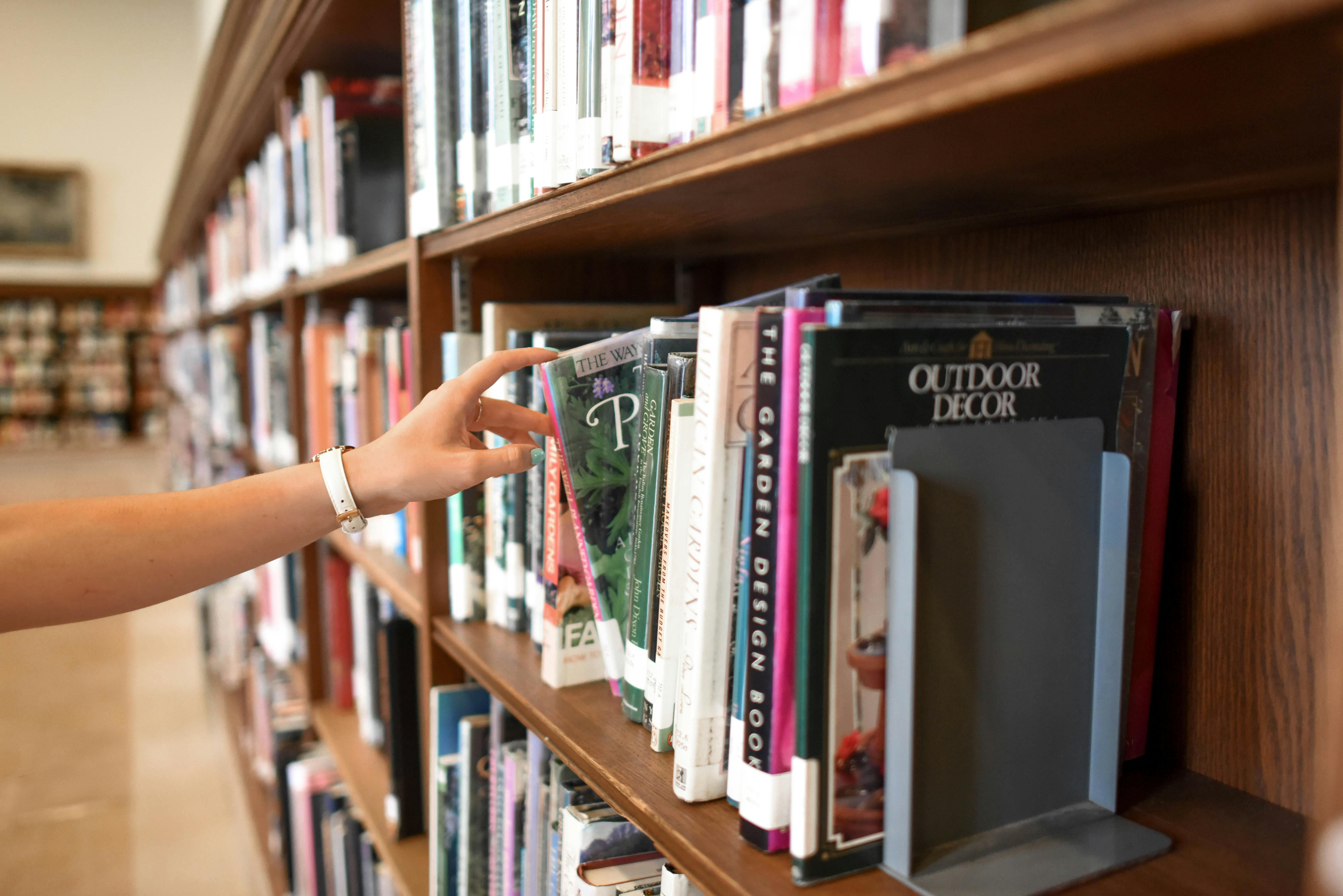 Hand reaching out to grab a book from a library shelf. The hand is shown mid-grasp, fingers extended towards the spine of a book. In the background are rows of neatly organized books on wooden shelves, creating a warm and inviting atmosphere. This is set as a real world example of how taxonomy and ontology enhance discoverability.