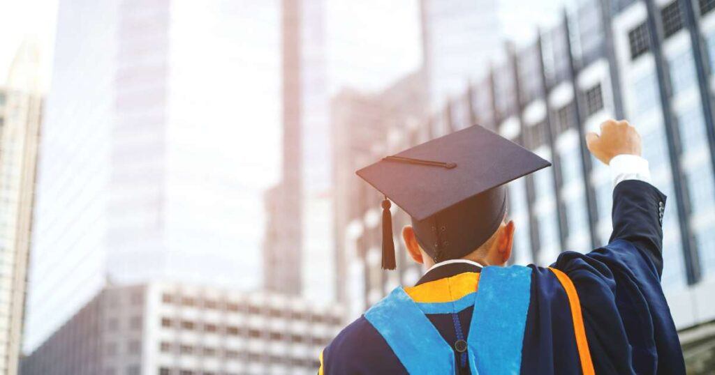 back of graduate's head in cap and gown fist pumping in front of city scape buildings