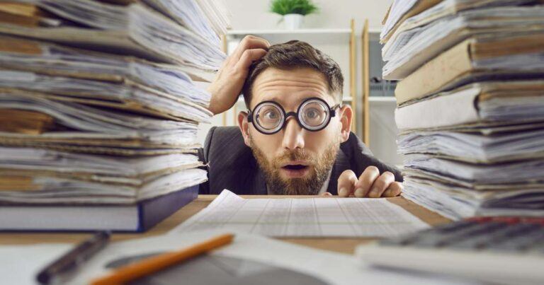 funny image of man peering at the camera with surprised look between two tall stacks of paperwork at his work desk