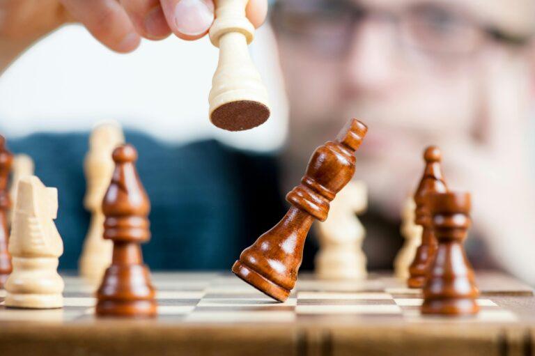 Chessboard with wooden pieces, one white piece lifted in mid-air by a hand, poised for a move. In focus, a brown king piece topples over, its base barely touching the board. Blurred background reveals a concentrated face with glasses, adding a strategic atmosphere. Competitive metaphor.