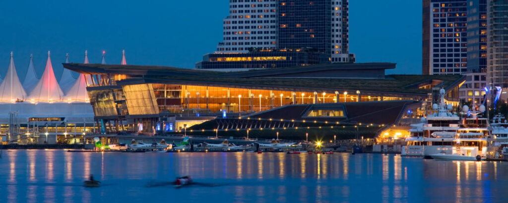 A photograph of the outside of the Vancouver Convention center taken in the evening. The building is lit from the outside. It is on the water.